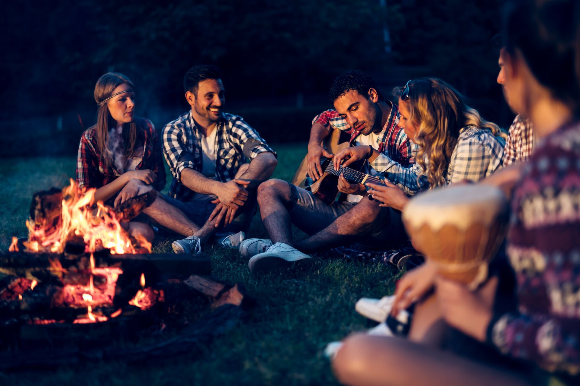 Group of friends around a campfire at night, with one playing guitar and another holding a drum.