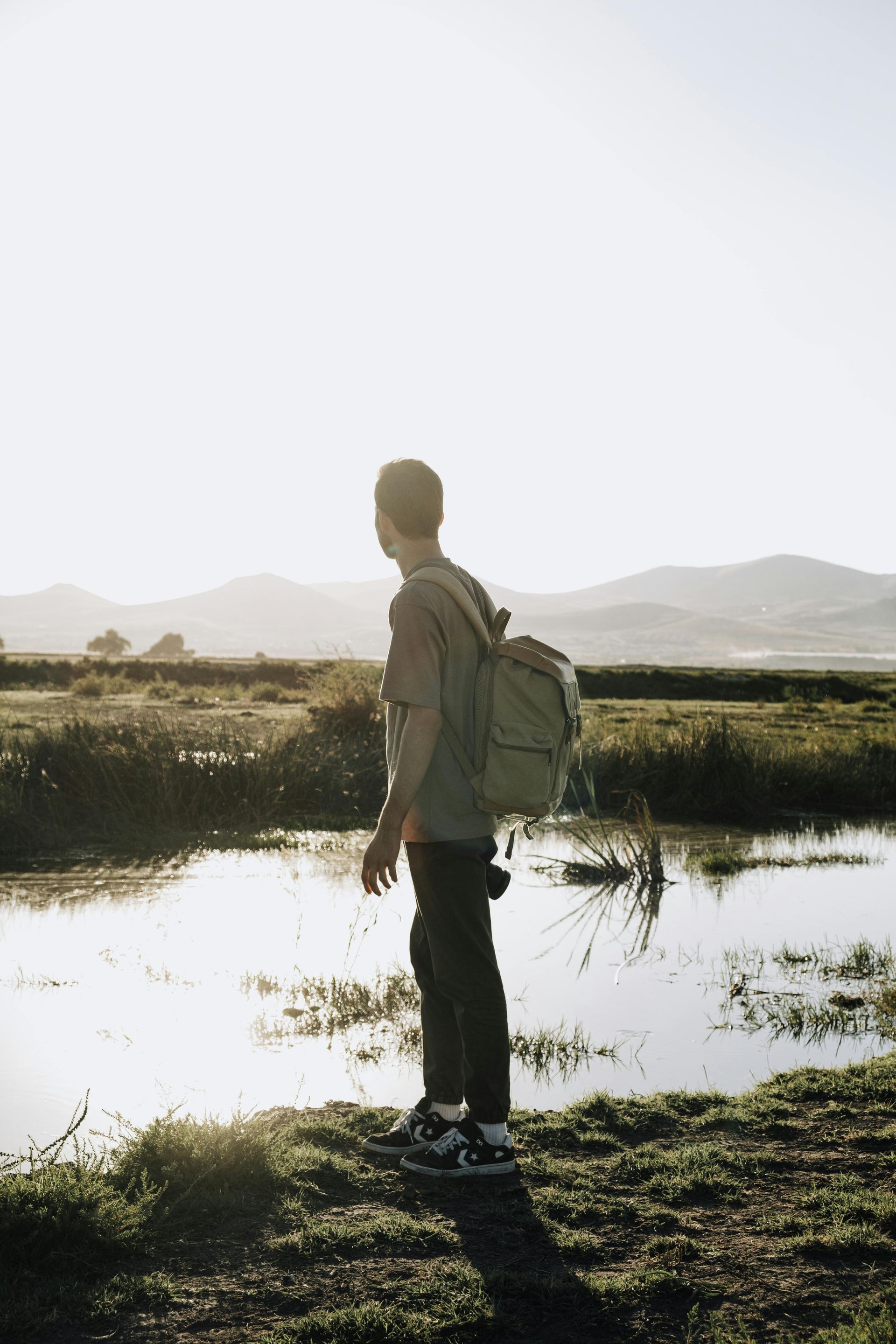 Person with backpack stands near water, looking toward distant mountains on a sunny day.