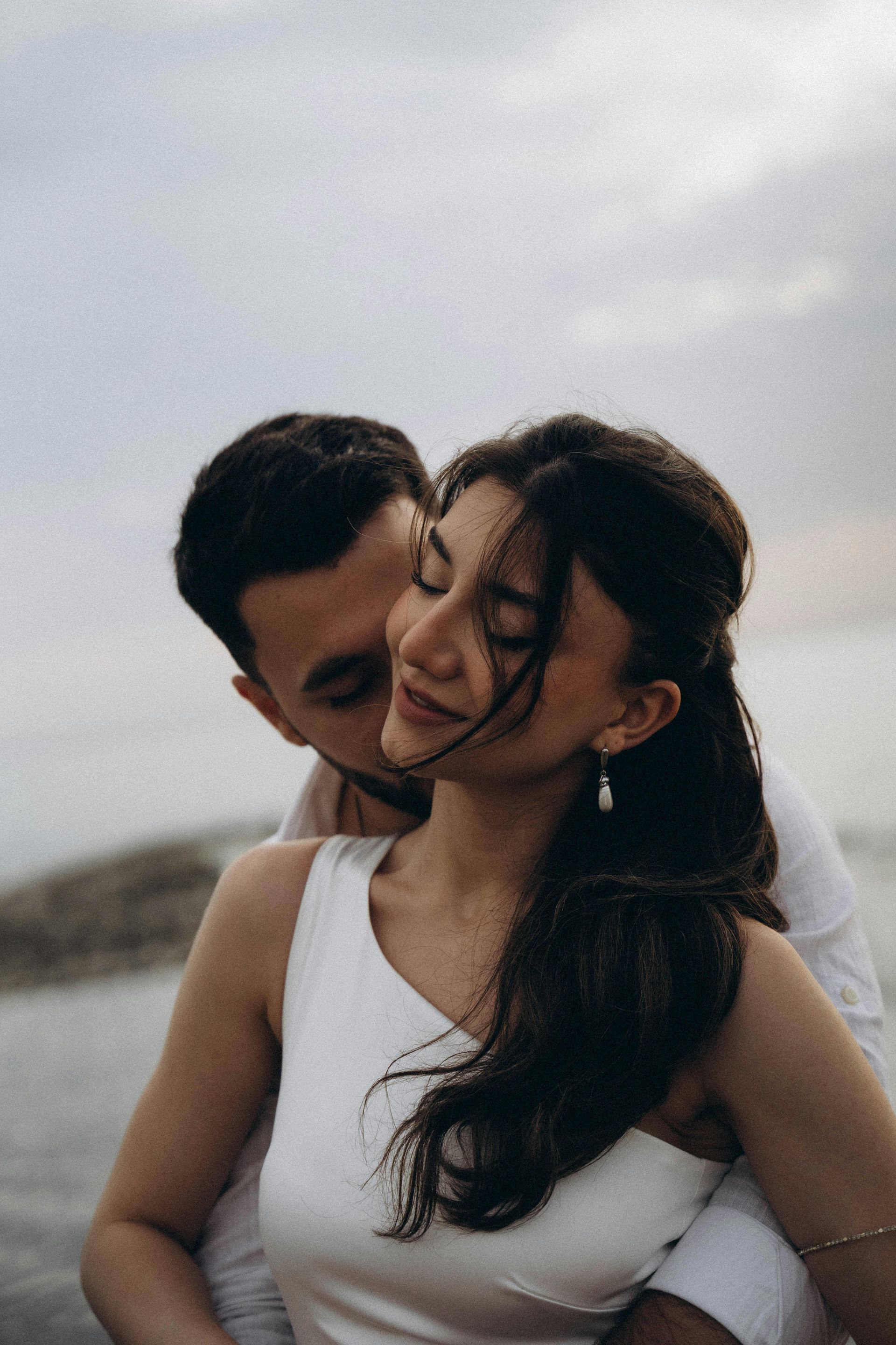 Man embracing woman, both smiling, outdoors; overcast sky.
