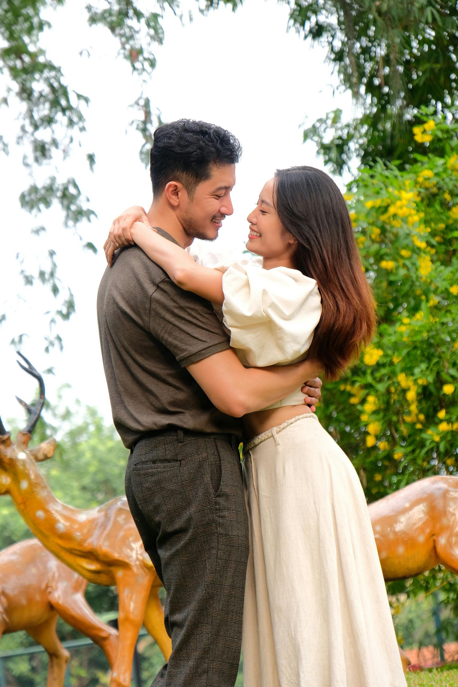 Couple hugging, smiling, in a park. Brown shirt and pants, white dress. Deer statues, trees, and flowers visible.