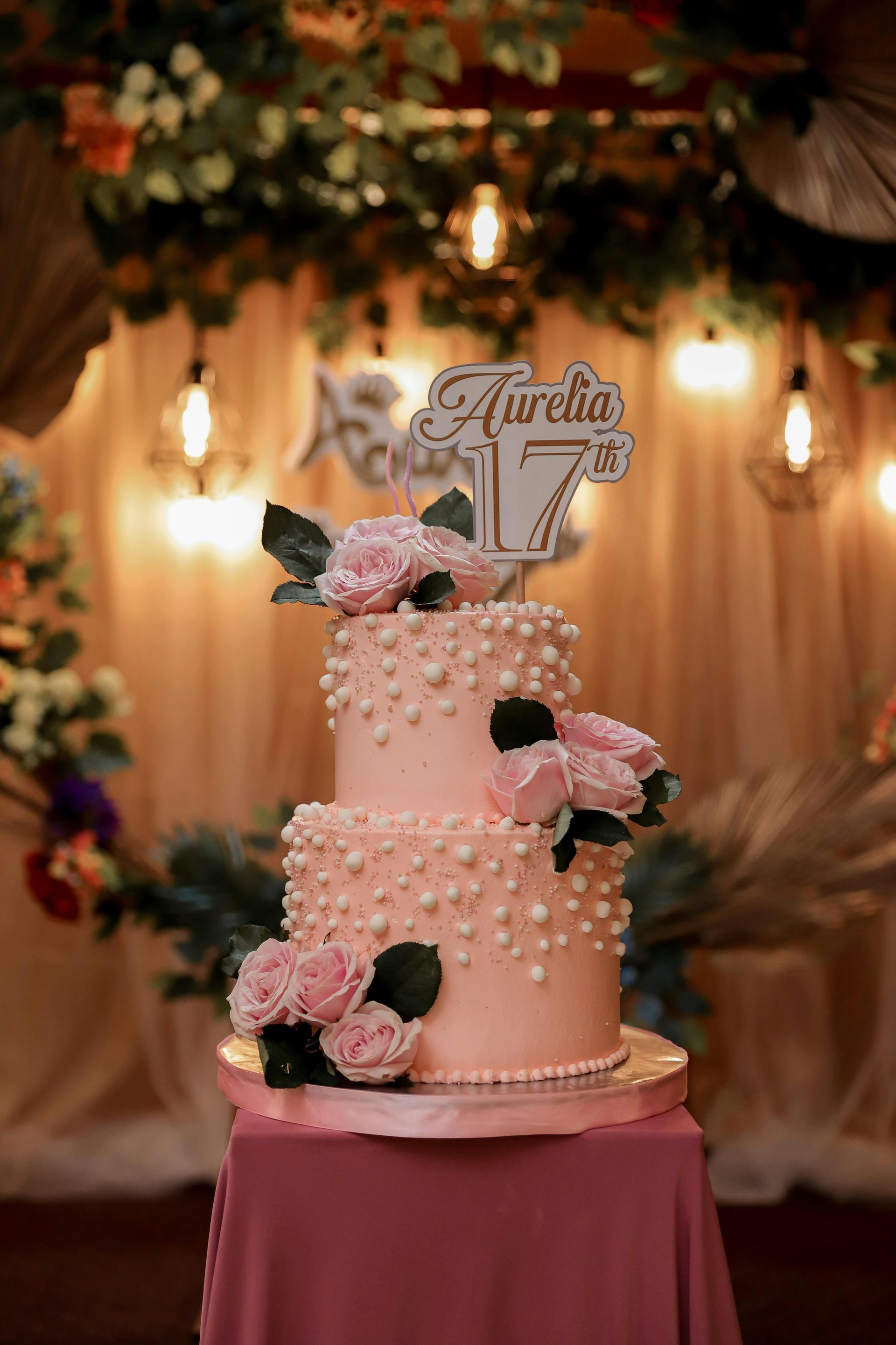 Two-tiered pink cake with pearls and roses, topped with a "17" and name, on a table. Decorated background.
