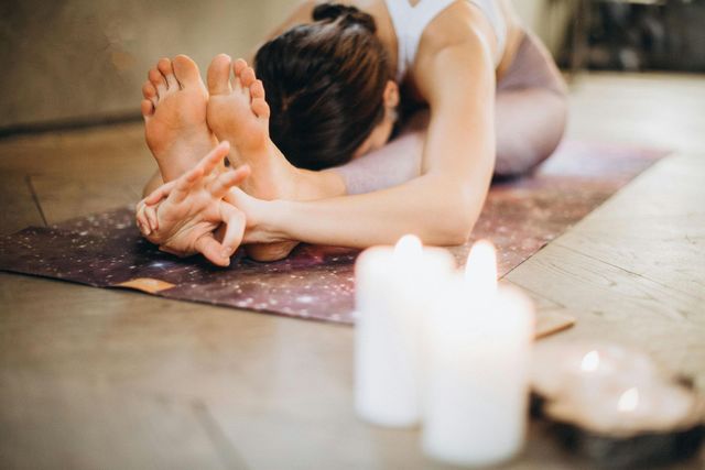 Woman in yoga pose, reaching for feet, on mat with candles nearby.