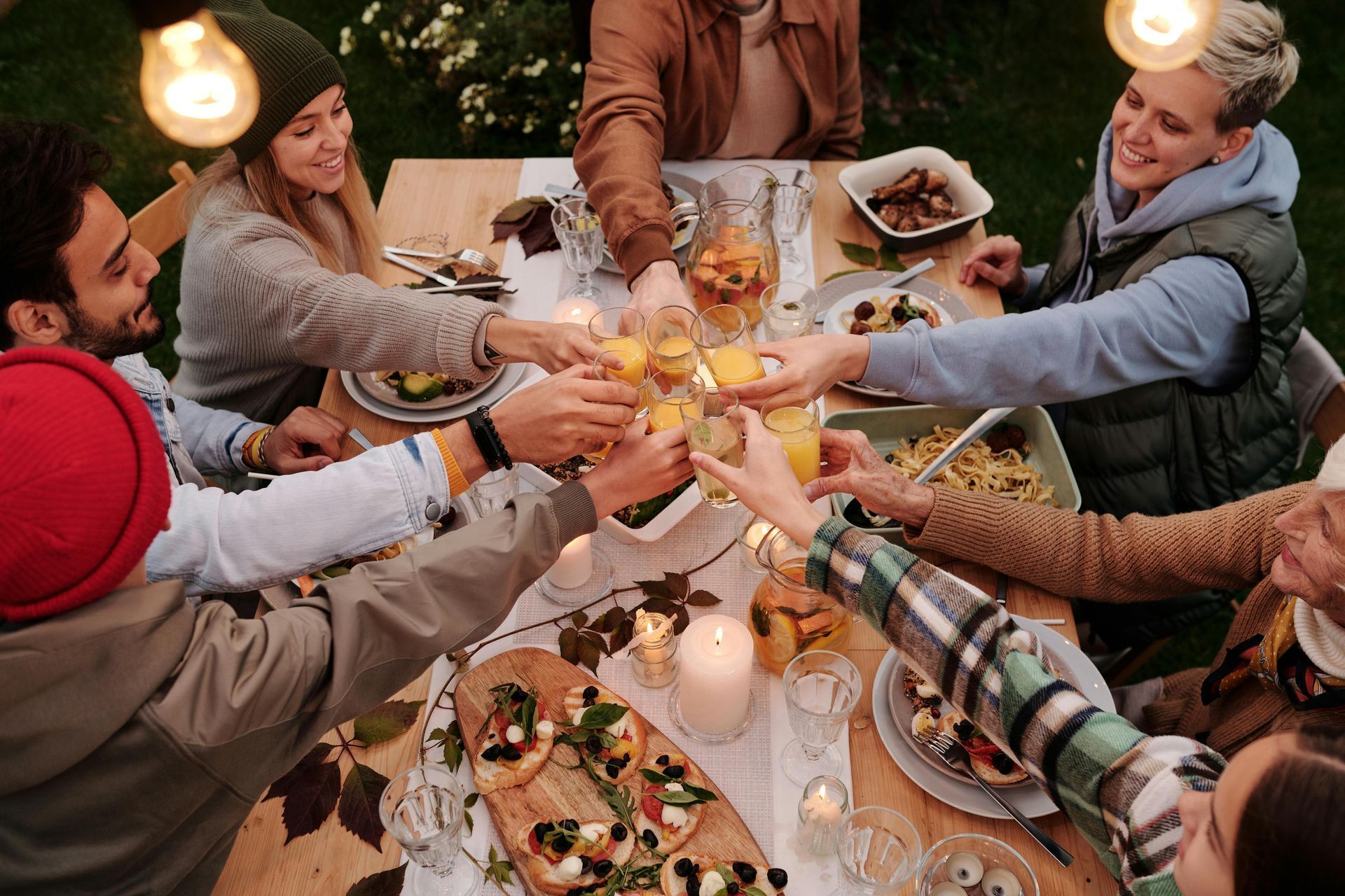 People toasting drinks around a wooden table set outdoors with food and candles, lit by hanging lights.