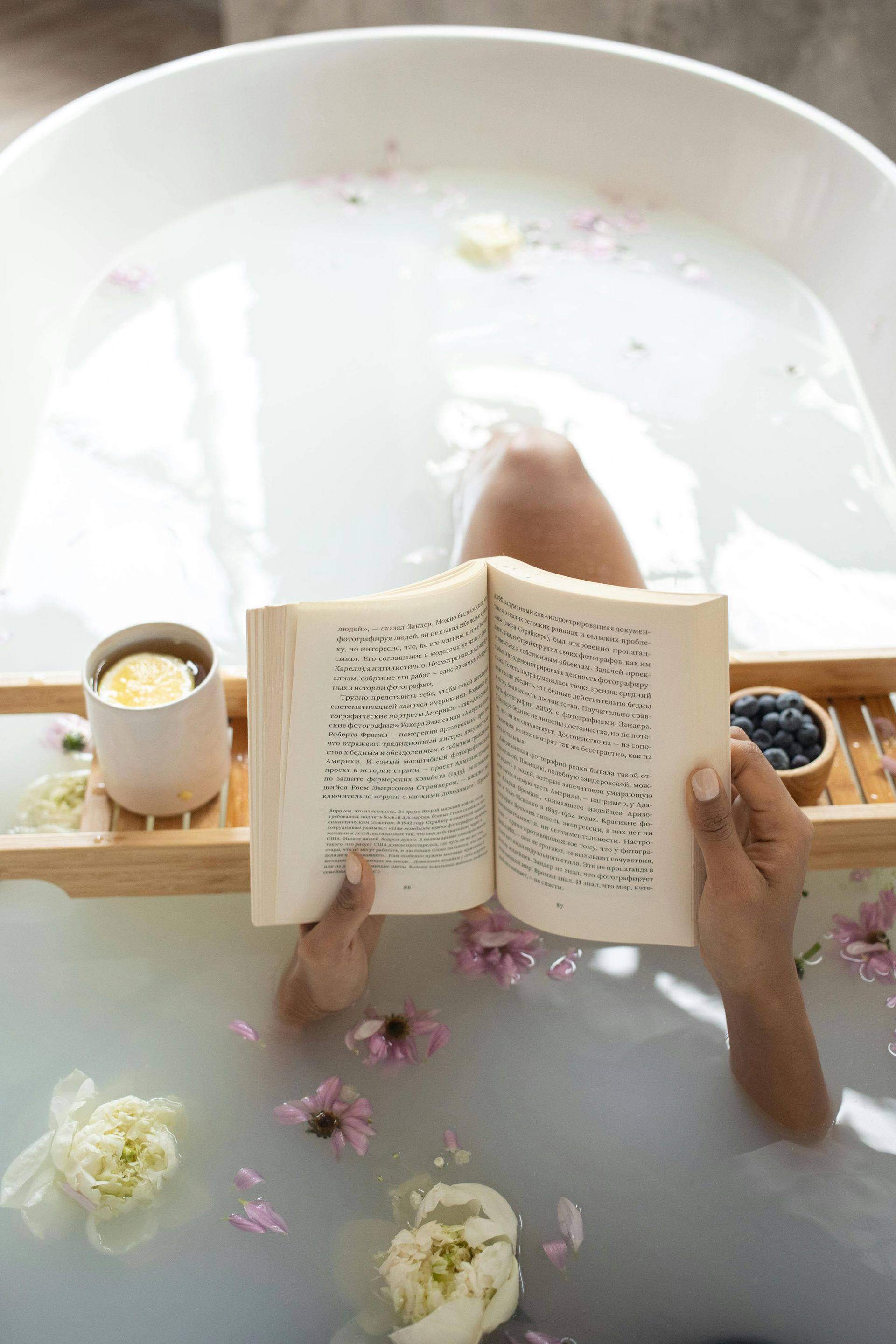 Person in a bathtub reading a book, wooden tray with a drink and blueberries, flower petals in the water.