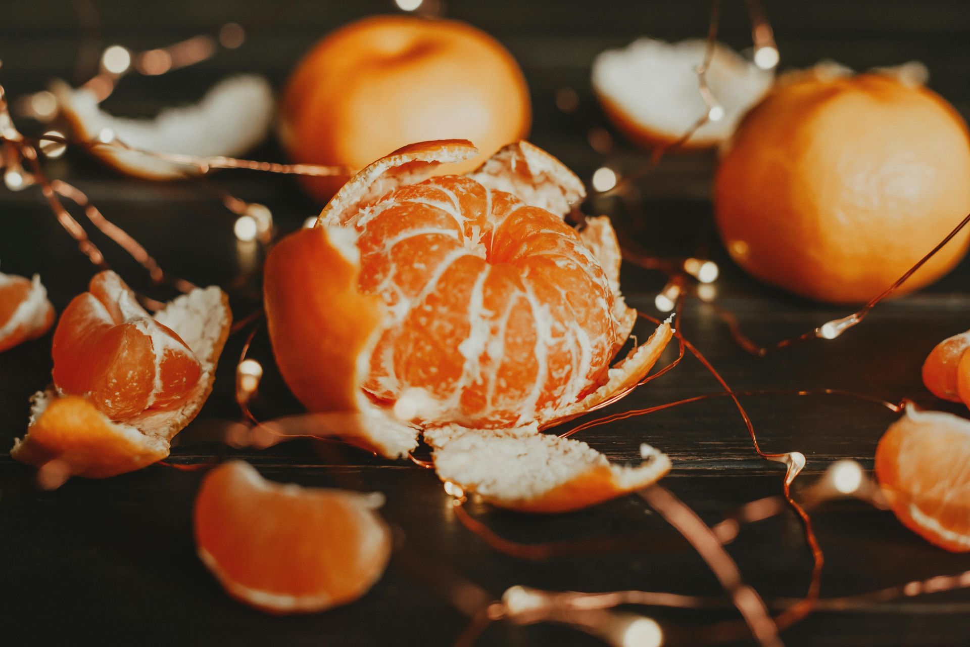 Close-up of peeled tangerines and segments with string lights on a dark surface.