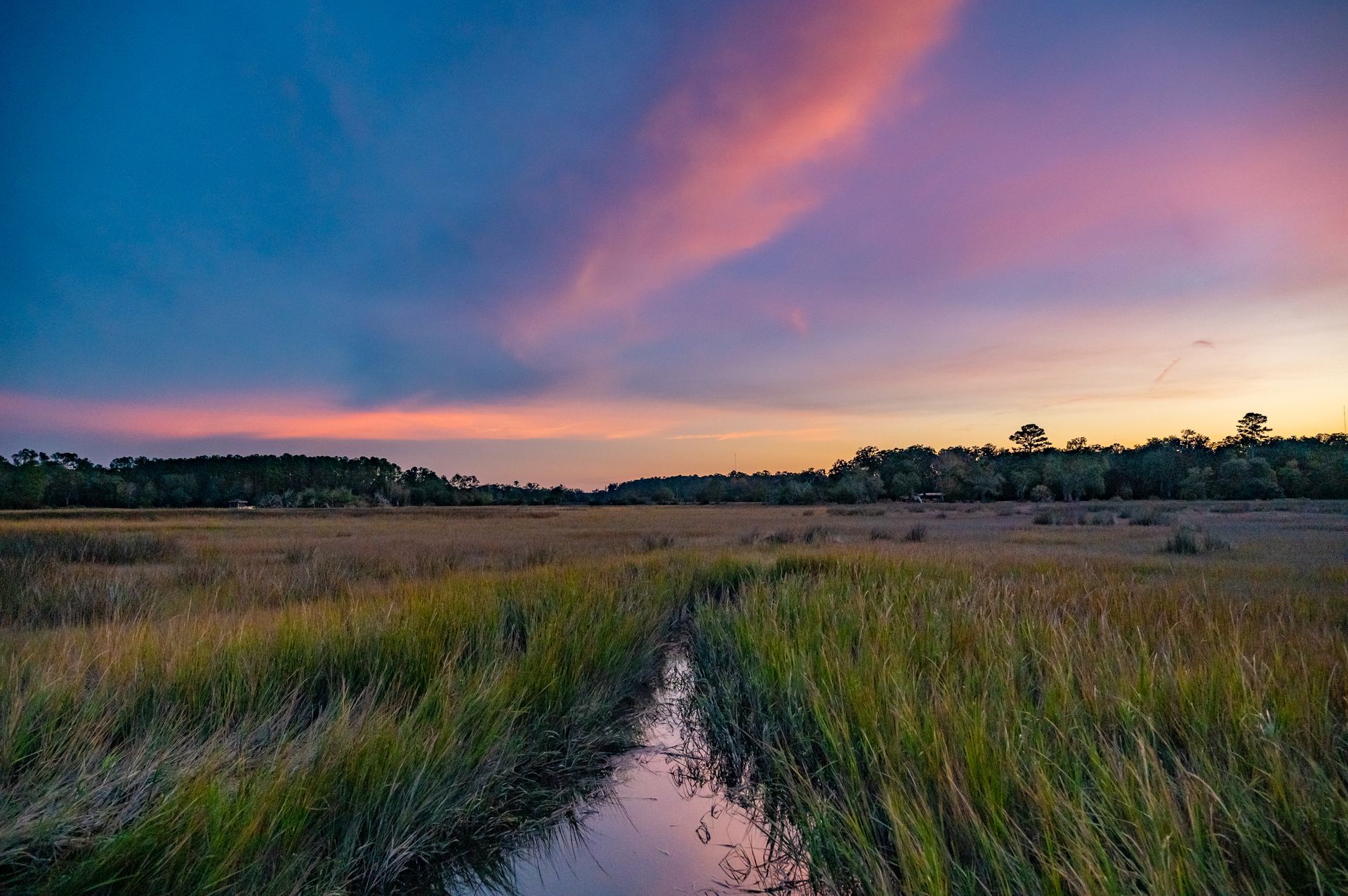 Sunset over a marsh with pink and blue sky; water channel through tall grasses.