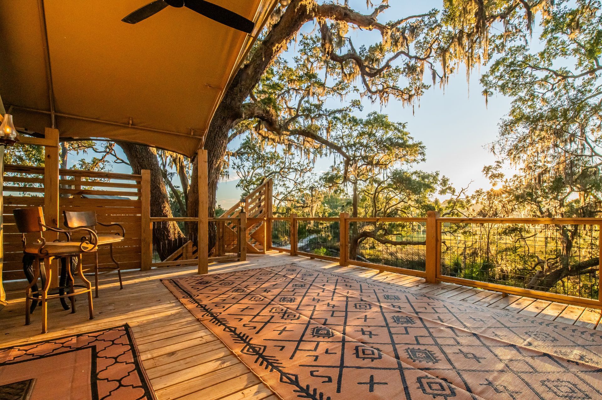 Wooden deck with rug, two chairs, and tree canopy in a sunny setting.