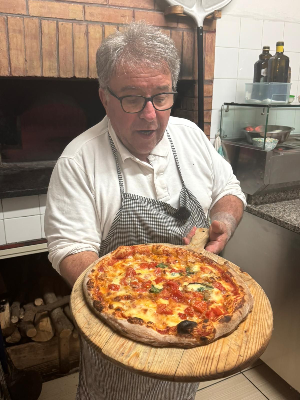 Uno chef tiene in mano una pizza appena sfornata su una pala di legno in una cucina con forno a legna.