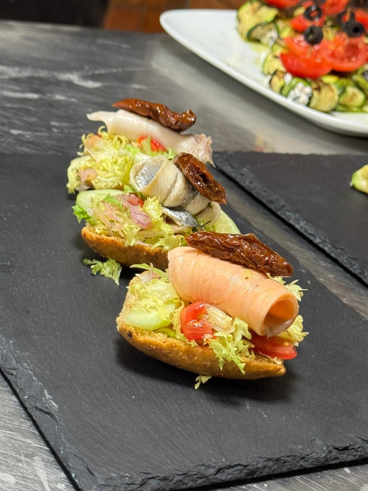 Tre tartine gourmet su un tagliere di ardesia, guarnite con salmone, verdure e pesce, su un tavolo scuro.
