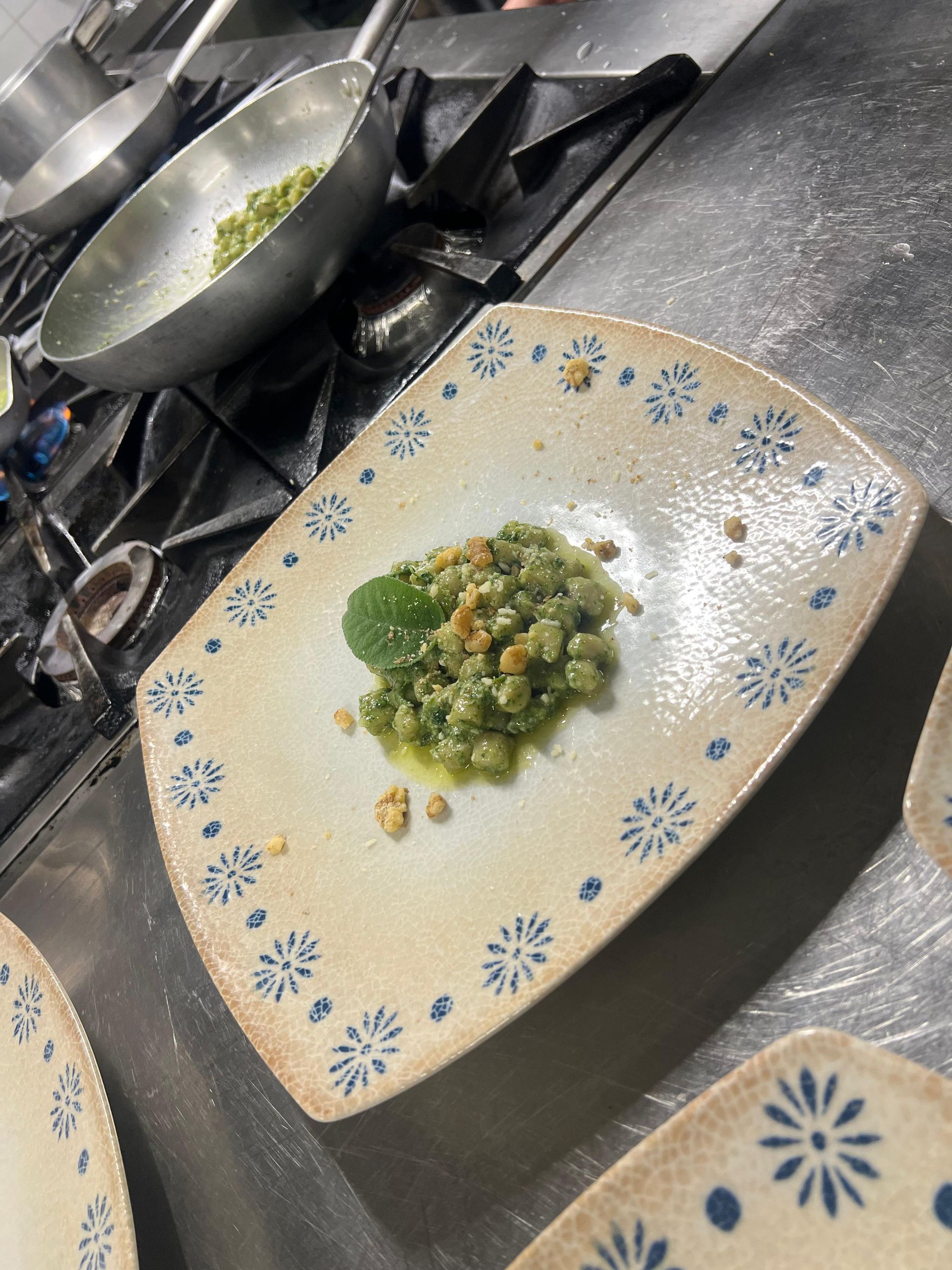 Un piatto bianco con fiori blu, su un piano cottura in acciaio inox, contiene una piccola ciotola di piselli verdi.