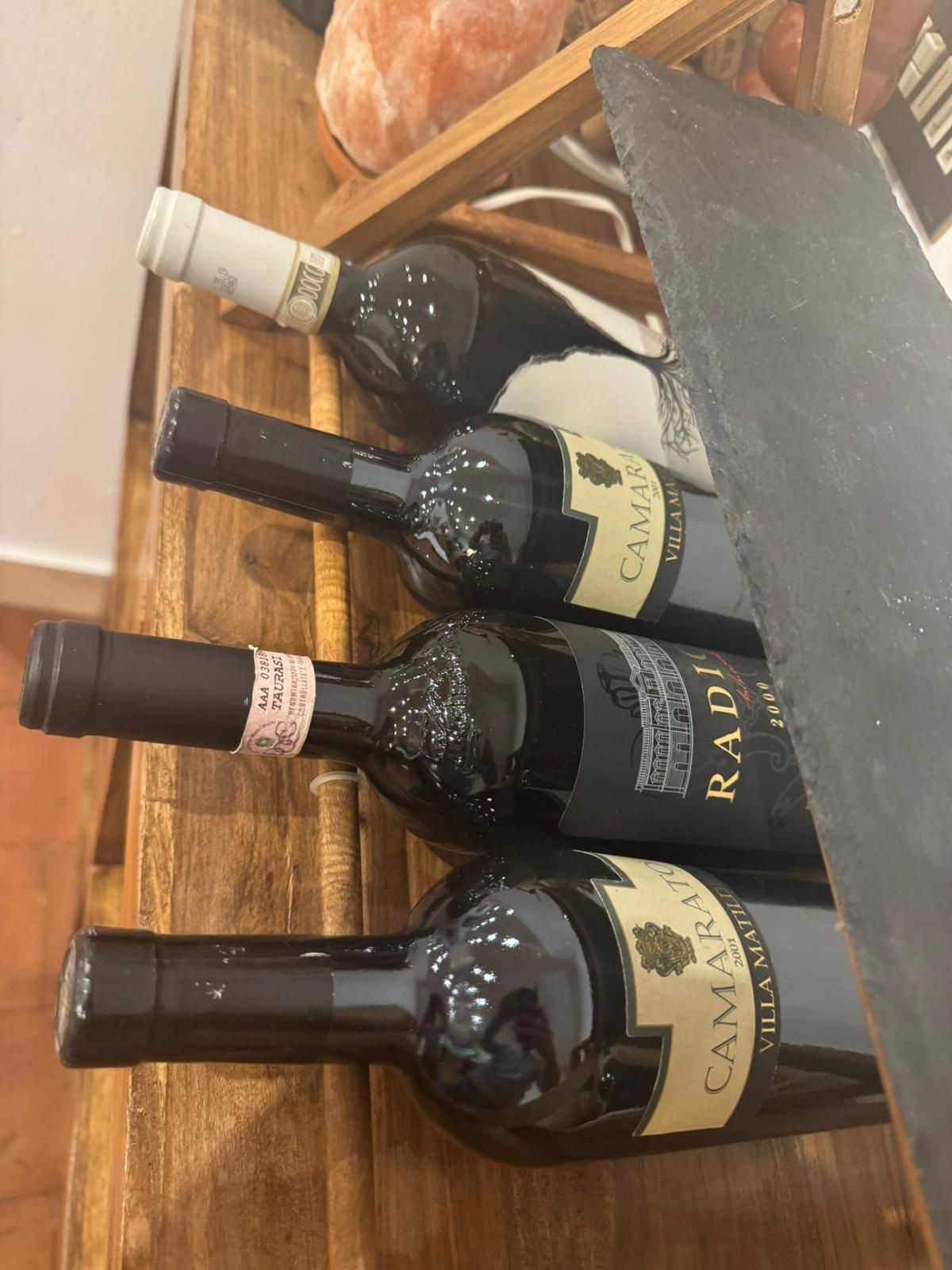 Quattro bottiglie di vino con etichette scure giacciono su un supporto di legno, viste di profilo.