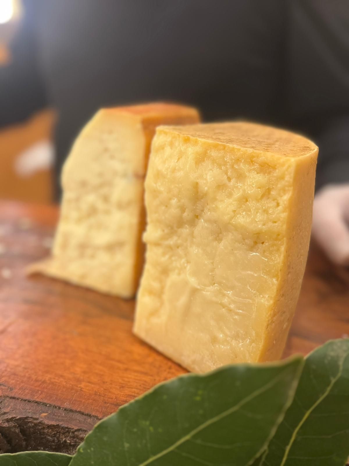 Due spicchi di formaggio giallo pallido su un tagliere di legno con una foglia verde in primo piano.