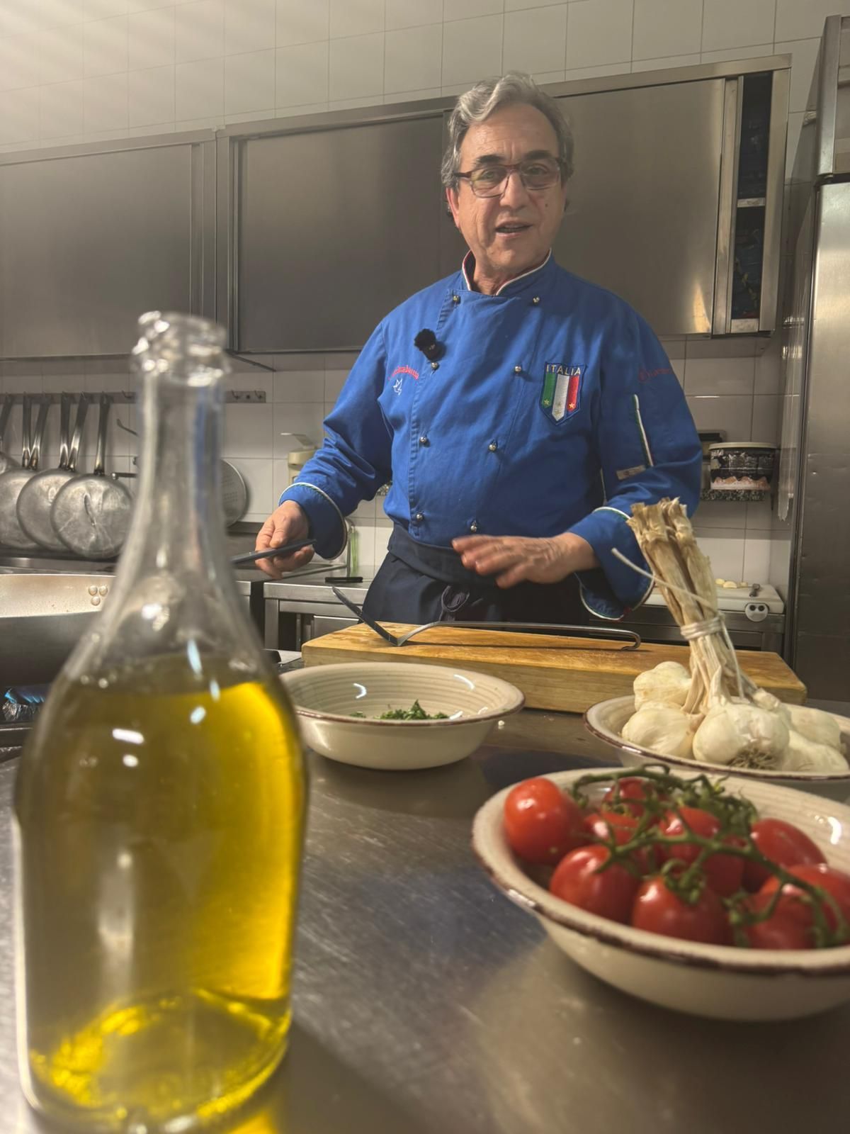 Uno chef in giacca blu prepara del cibo in cucina, con olio d'oliva e ciotole di pomodori e aglio in primo piano.