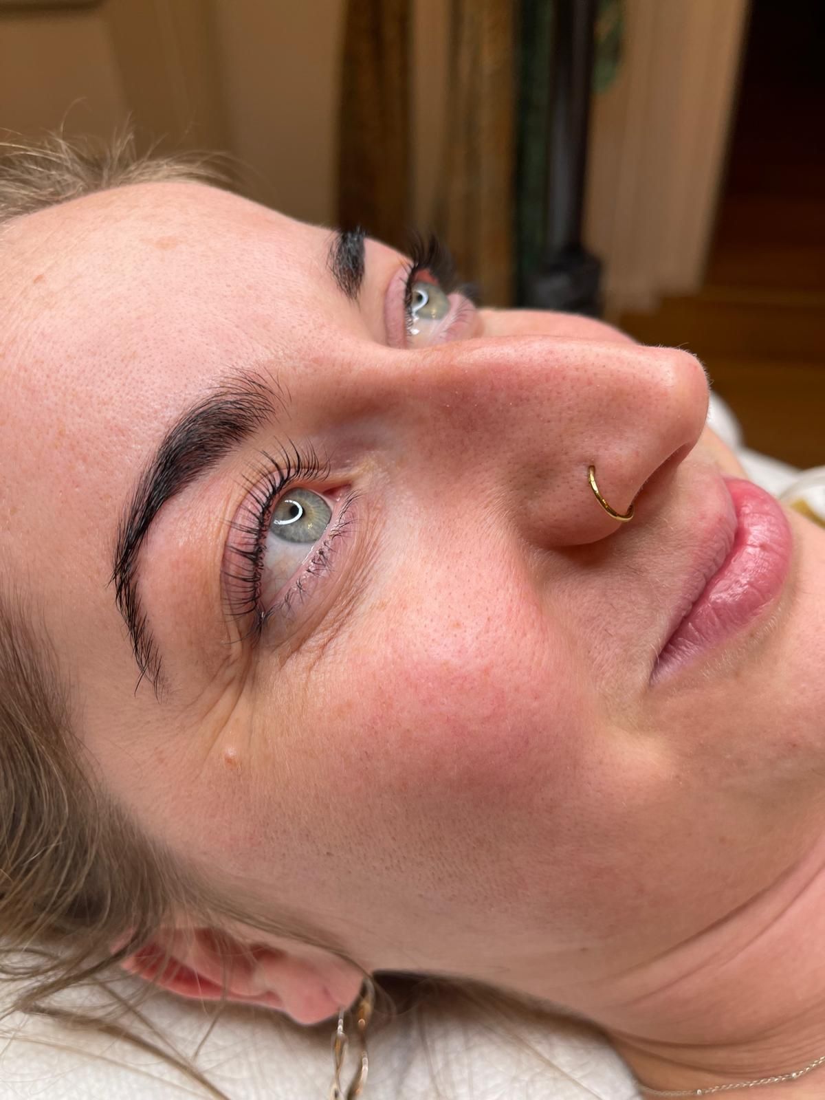 Nahaufnahme einer Person, die auf einem Bett liegt, nach oben schaut und einen Nasenring sowie natürliches Make-up trägt.