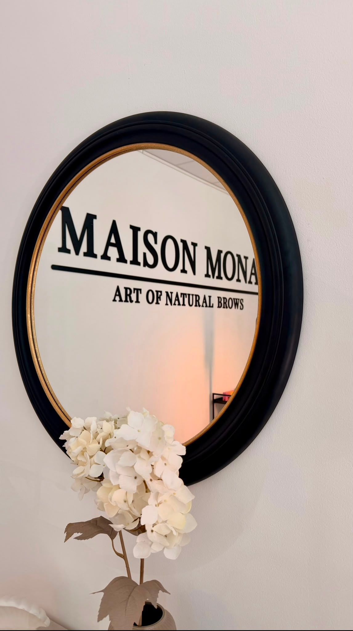 Runder, schwarz gerahmter Spiegel mit dem Schriftzug „MAISON MONA“ und weißen Blumen darunter an einer beigen Wand