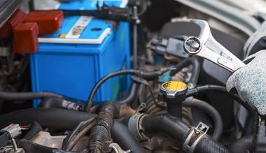 Mechanic working on car engine, holding wrench; blue battery visible.