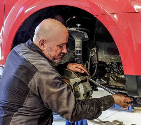 A mechanic uses a long metal tool to work on the brake assembly inside the wheel well of a red car.