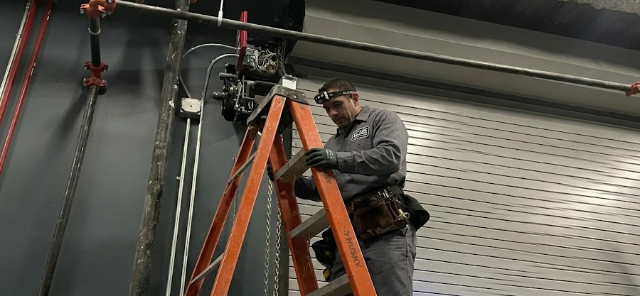 A person in work clothes standing on a ladder while repairing machinery mounted on a grey wall in a warehouse setting.