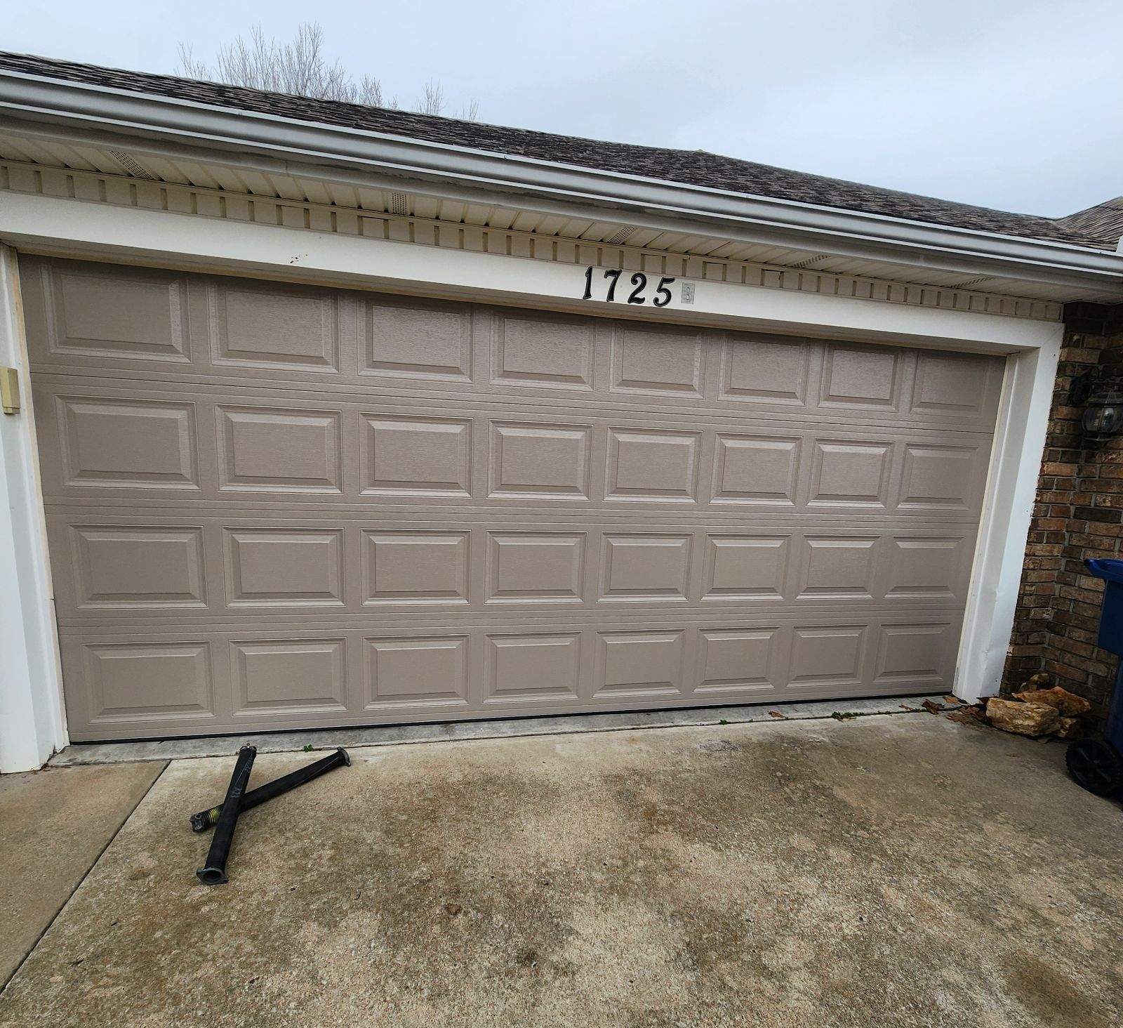 A tan, multi-paneled residential garage door with the number 1725 above it and a metal object lying on the concrete drive.