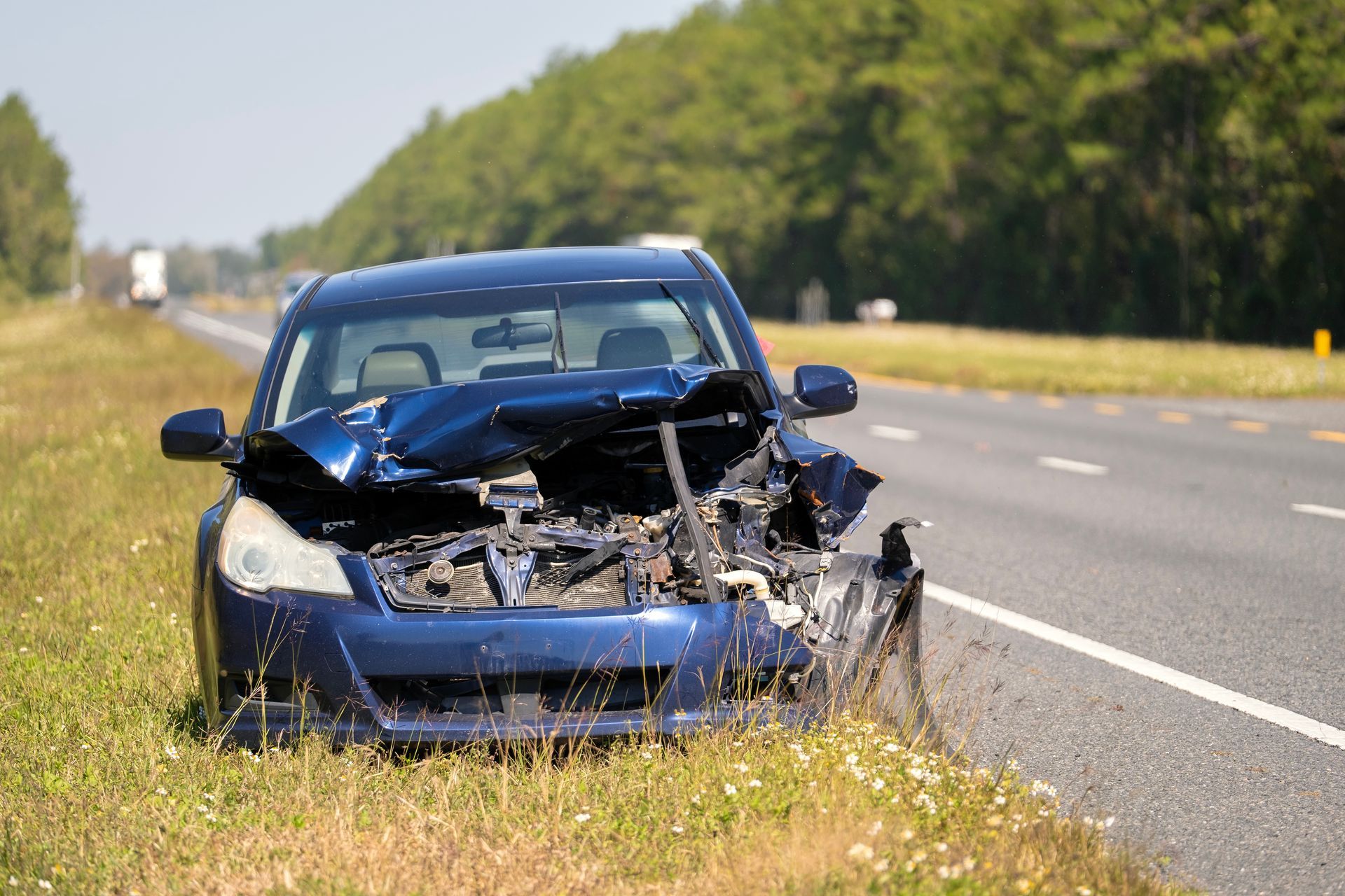 Blue sedan with a badly crushed front end sitting in roadside grass, waiting for junk car removal.