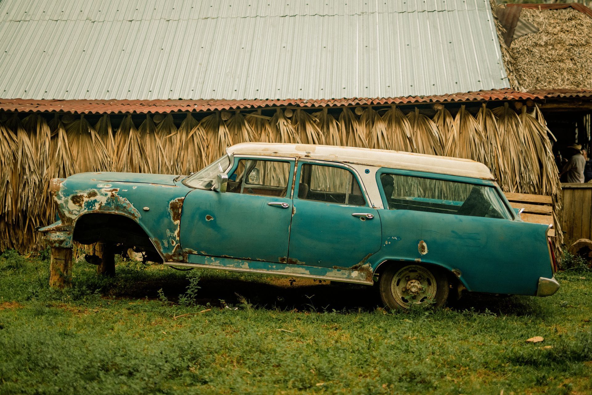 A rusty teal vintage station wagon propped up on blocks in front of a thatched-roof barn. A rusty teal vintage station wagon propped up on blocks in front of a thatched-roof barn.