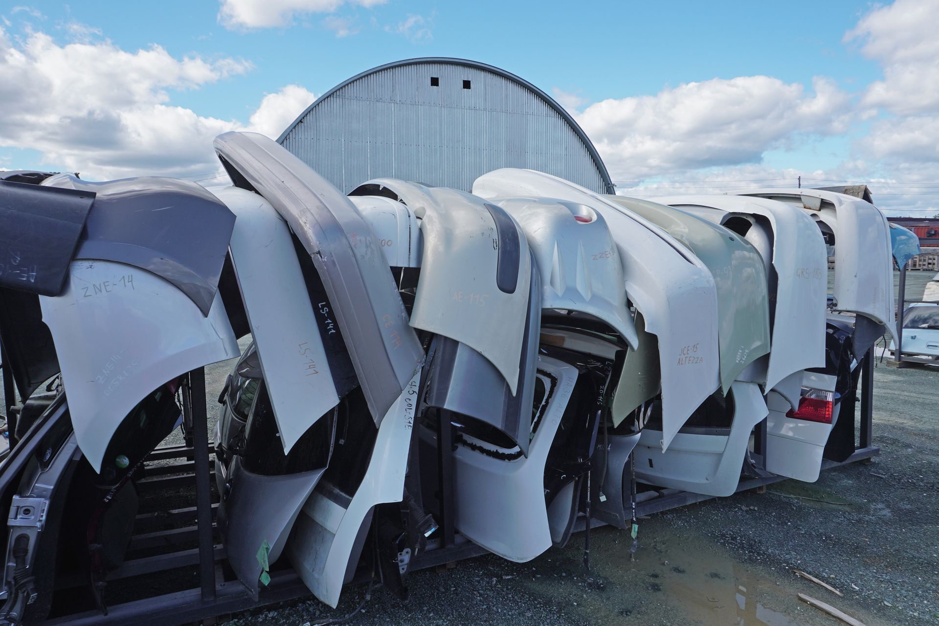 Salvaged auto hoods and fenders stacked outdoors in a local junk car buyer's yard under blue skies.