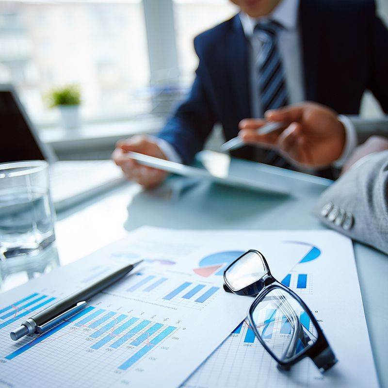Various data sheets on a table with businessmen looking at a tablet.