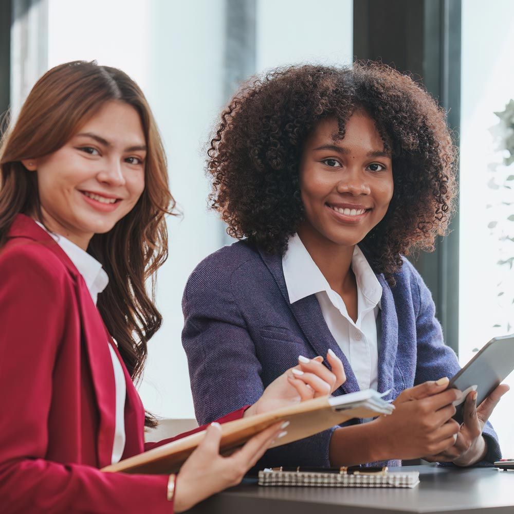 two women are sitting at a table looking at a tablet and other notes