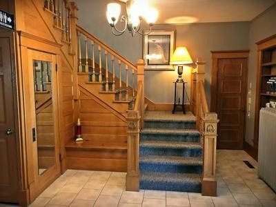 A wooden staircase with blue carpet leads upward in a foyer featuring a hanging chandelier, a floor lamp, and tile floors.
