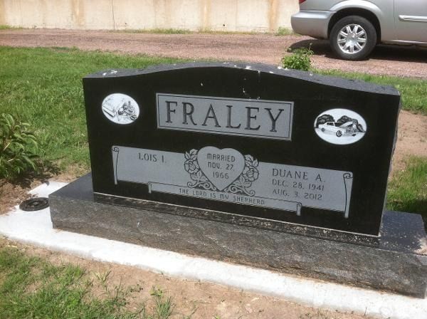 Black granite gravestone for the Fraley family in a grassy outdoor setting, showing dates and decorative car etchings.