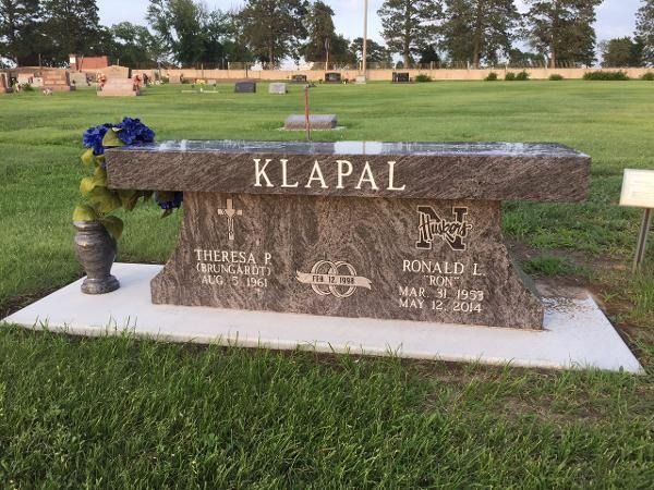 A granite bench gravestone for the Klapal family in a cemetery, featuring names, dates, and a decorative floral vase.