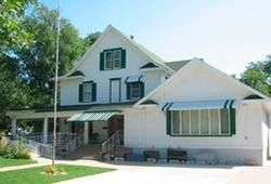 Two-story white building with green shutters and awnings, featuring a wrap-around porch and a metal accessibility ramp.