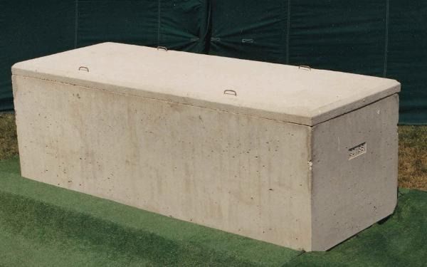 A beige, rectangular concrete burial vault sits on a patch of artificial grass in front of a green privacy screen.