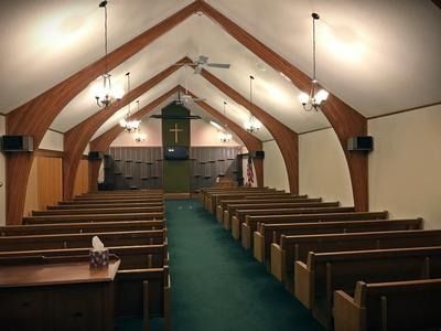 An interior view of a church sanctuary with wooden arched ceiling beams, rows of wooden pews, and a green carpeted aisle.