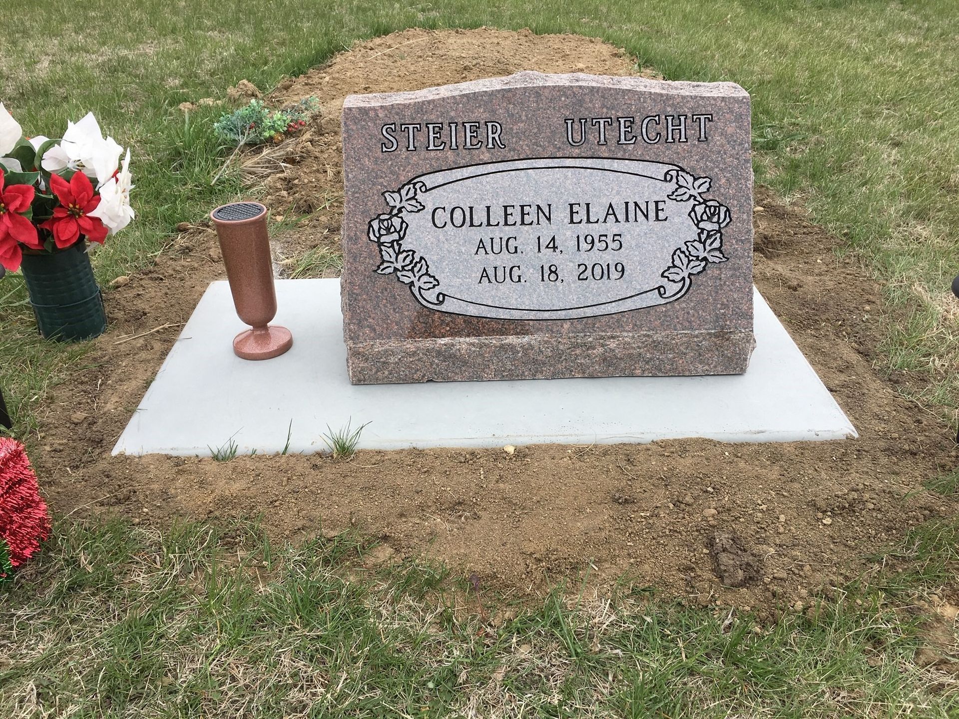 A red granite headstone for Colleen Elaine, marked 1955–2019, sits on a concrete base in a cemetery with floral tributes.