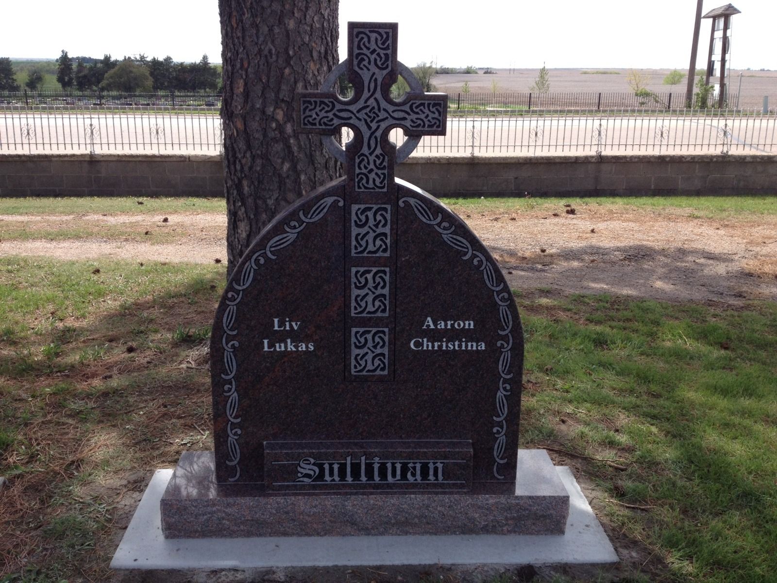A polished granite Sullivan family headstone with a Celtic cross, set in a cemetery before a tree.