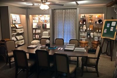A funeral planning room with a conference table, chairs, and shelves displaying urns, keepsakes, and printed materials.