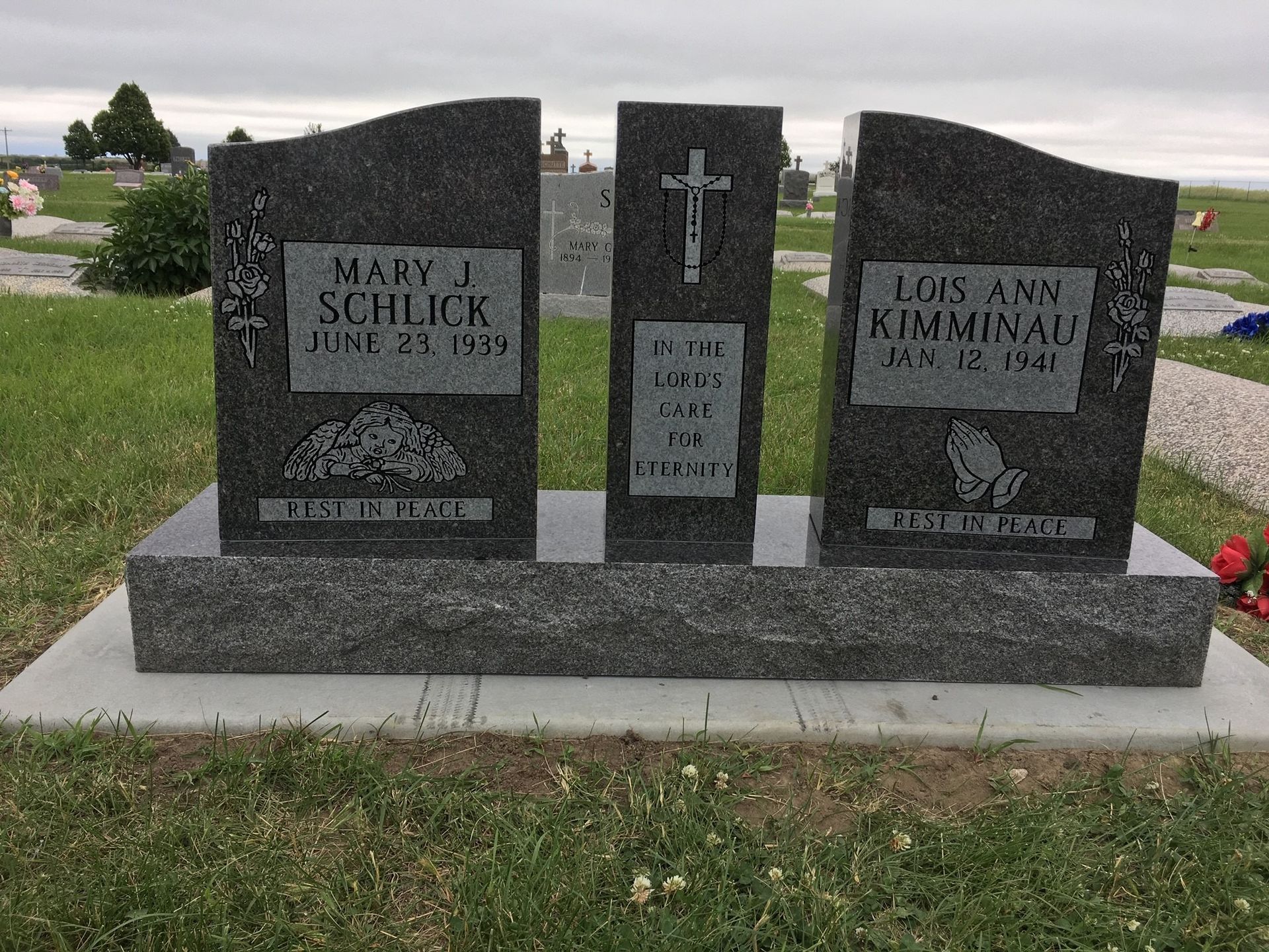 A dark grey granite headstone in a cemetery with a central cross and two panels, one for Mary J. Schlick and Lois Ann Kirmay.