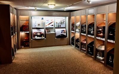 A funeral home showroom featuring rows of casket displays in lit wall compartments with neutral-toned walls and carpet.