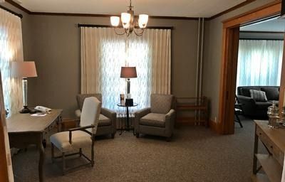 A well-lit office with a desk, office chair, two armchairs, side table with a lamp, and view into an adjacent lounge.