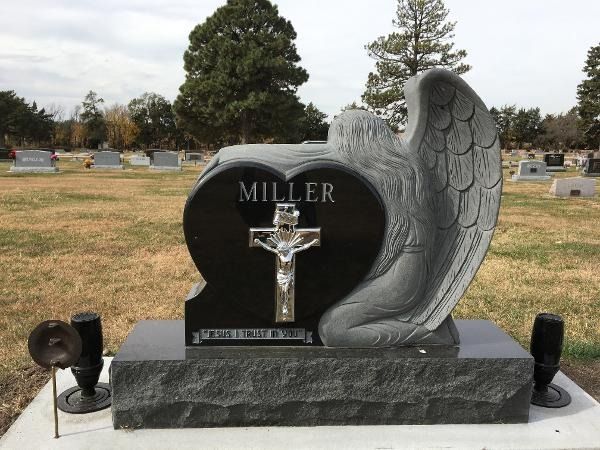 A black heart-shaped headstone featuring a metallic crucifix and a stone angel, set in a grassy cemetery.