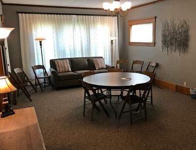 A round table with folding chairs, a sofa by a large window with sheer curtains, and lamps in a grey-walled room.