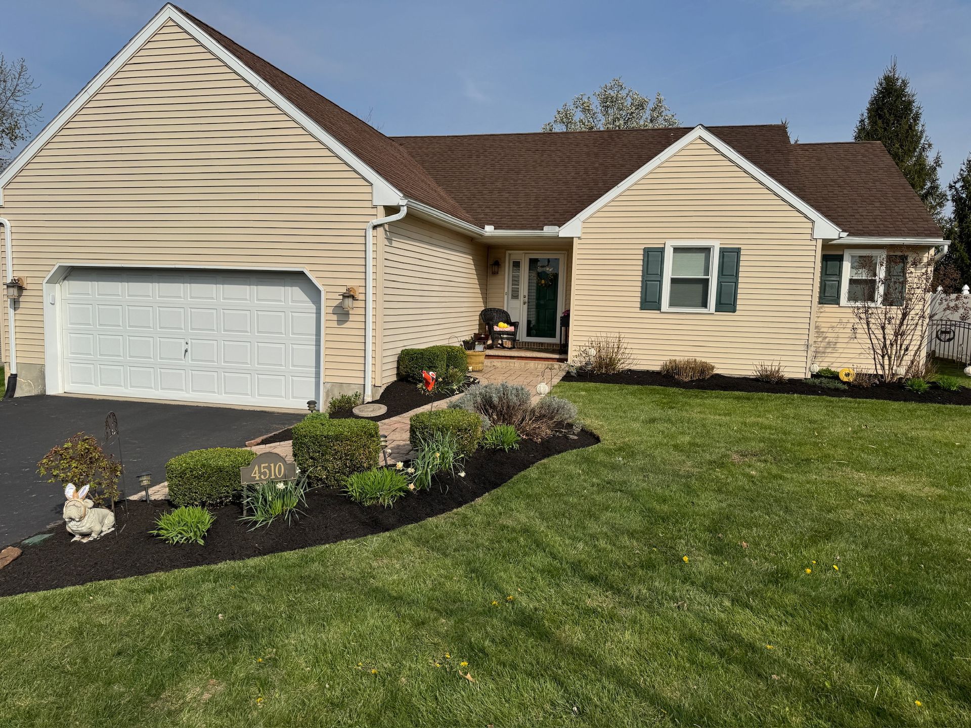Tan house with white garage door, green shutters, and well-kept lawn with black mulch flowerbeds.