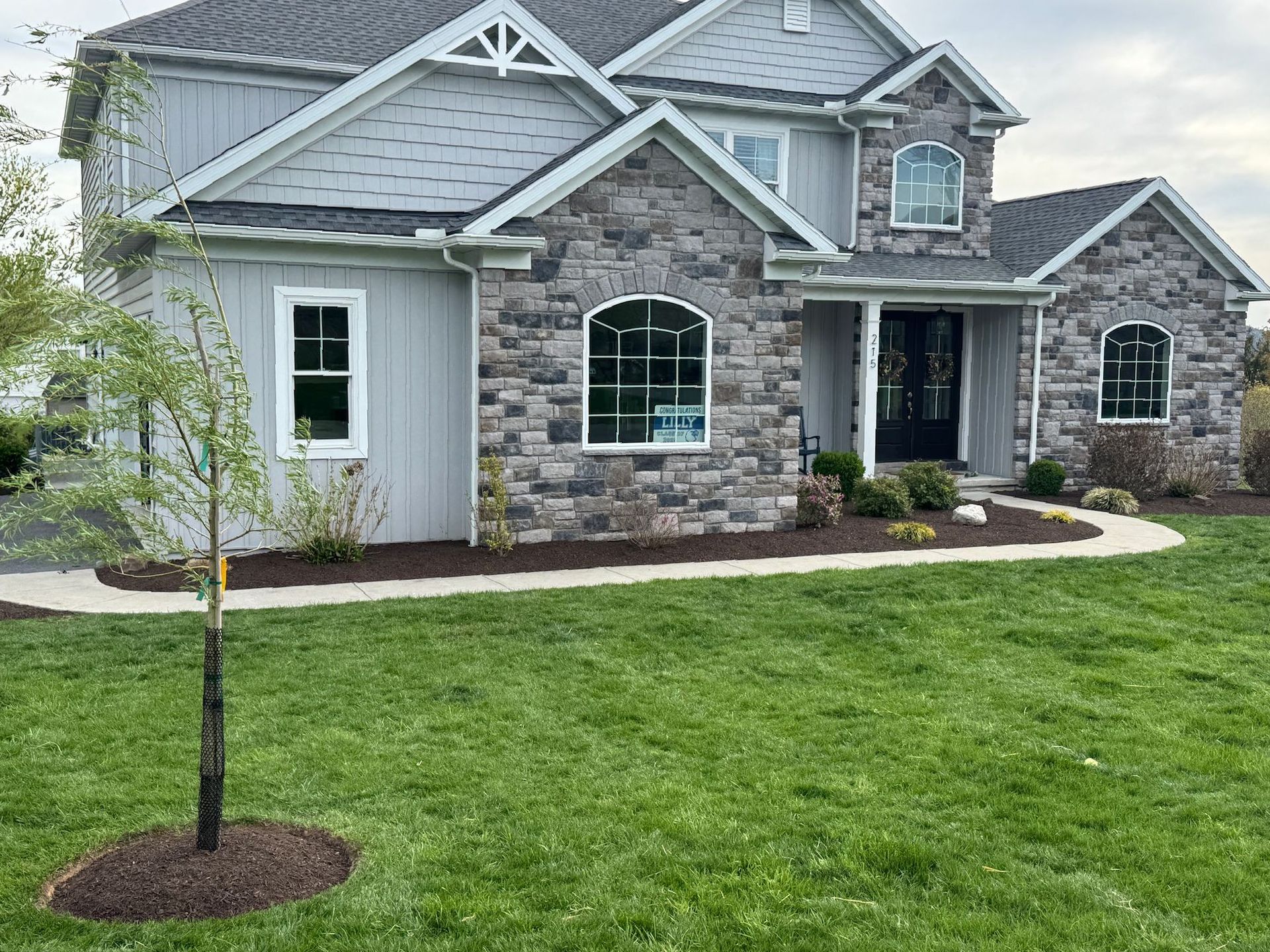Two-story house with stone facade, gray siding, and arched windows, surrounded by green grass and landscaping.