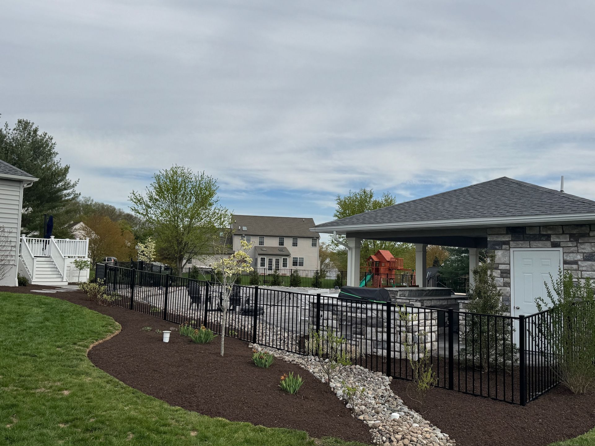 Backyard with black fence, stone-covered bar, and a pool. Green lawn, trees, and cloudy sky.