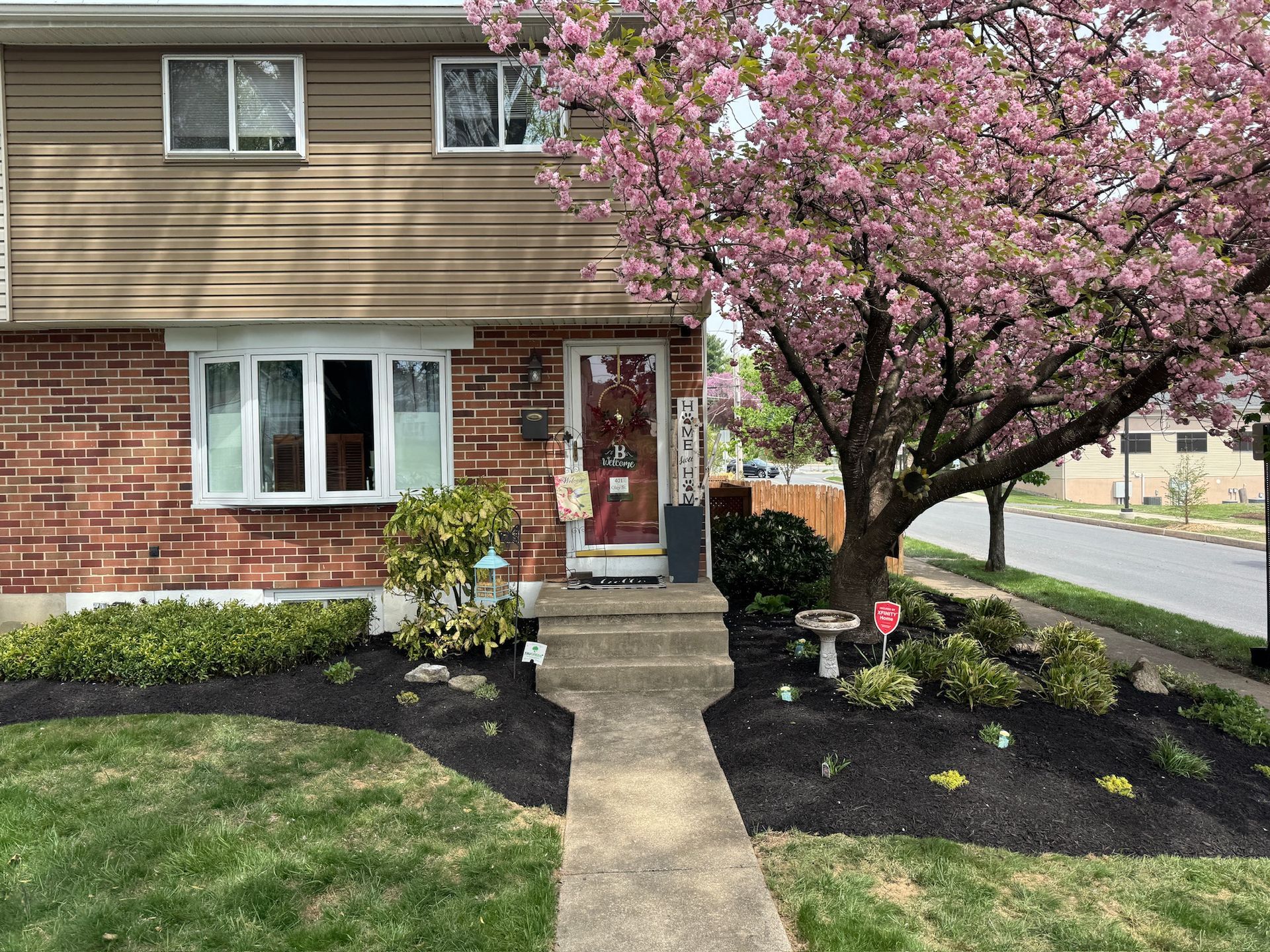 A brick and wood-paneled townhouse with a blossoming pink tree in front.  A concrete walkway leads to the front door.