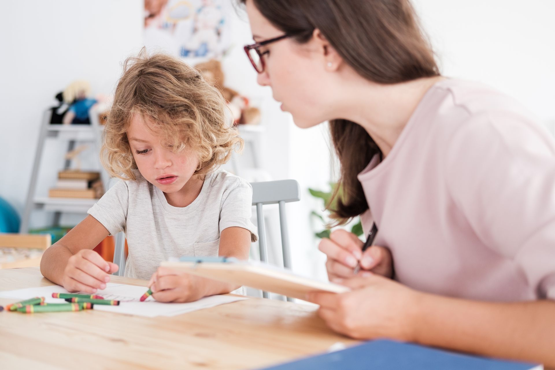 Child drawing at a table with an adult. The adult, wearing glasses, points at paper. Crayons on table.