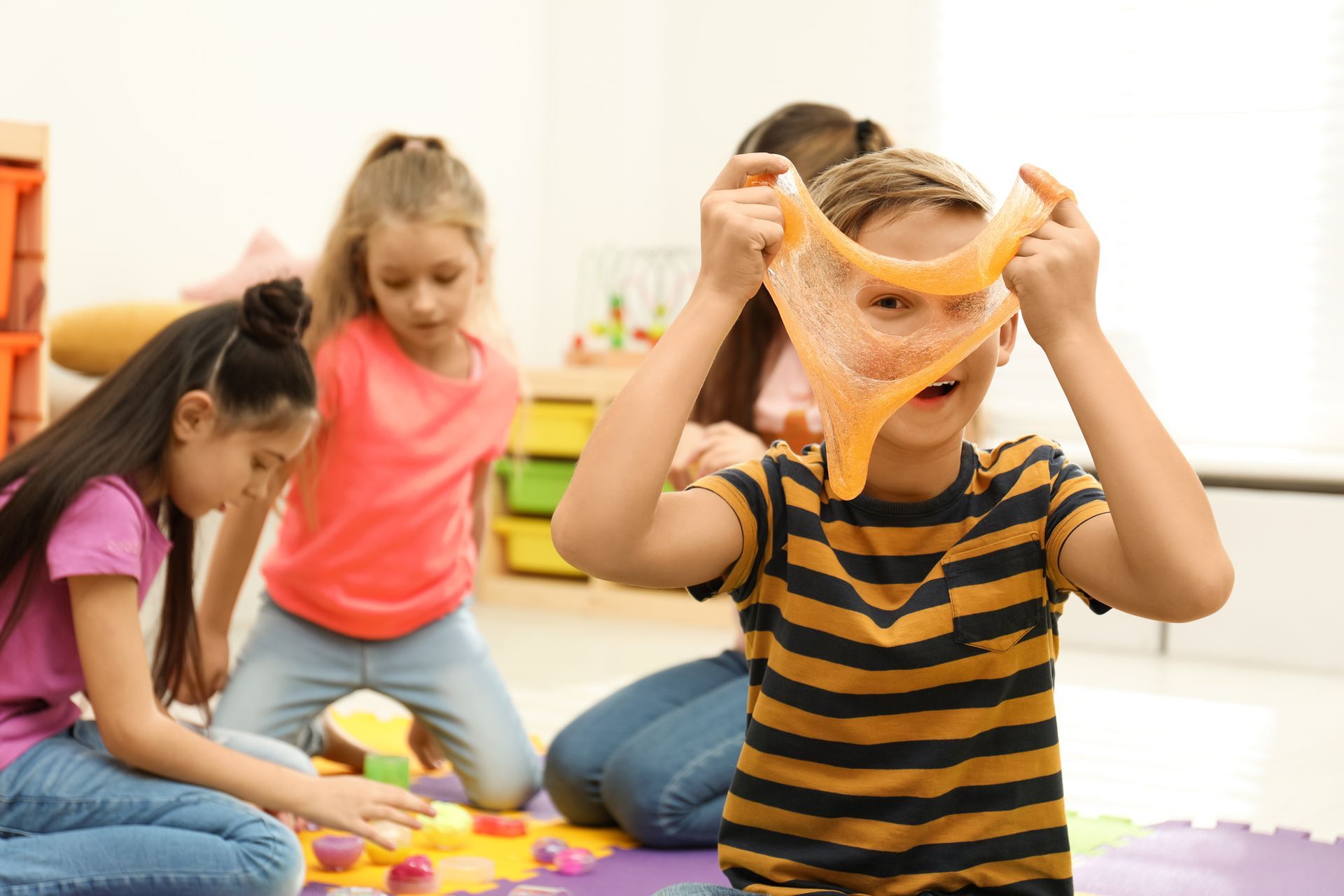 Boy holding orange slime, surrounded by other children playing with slime in a classroom.