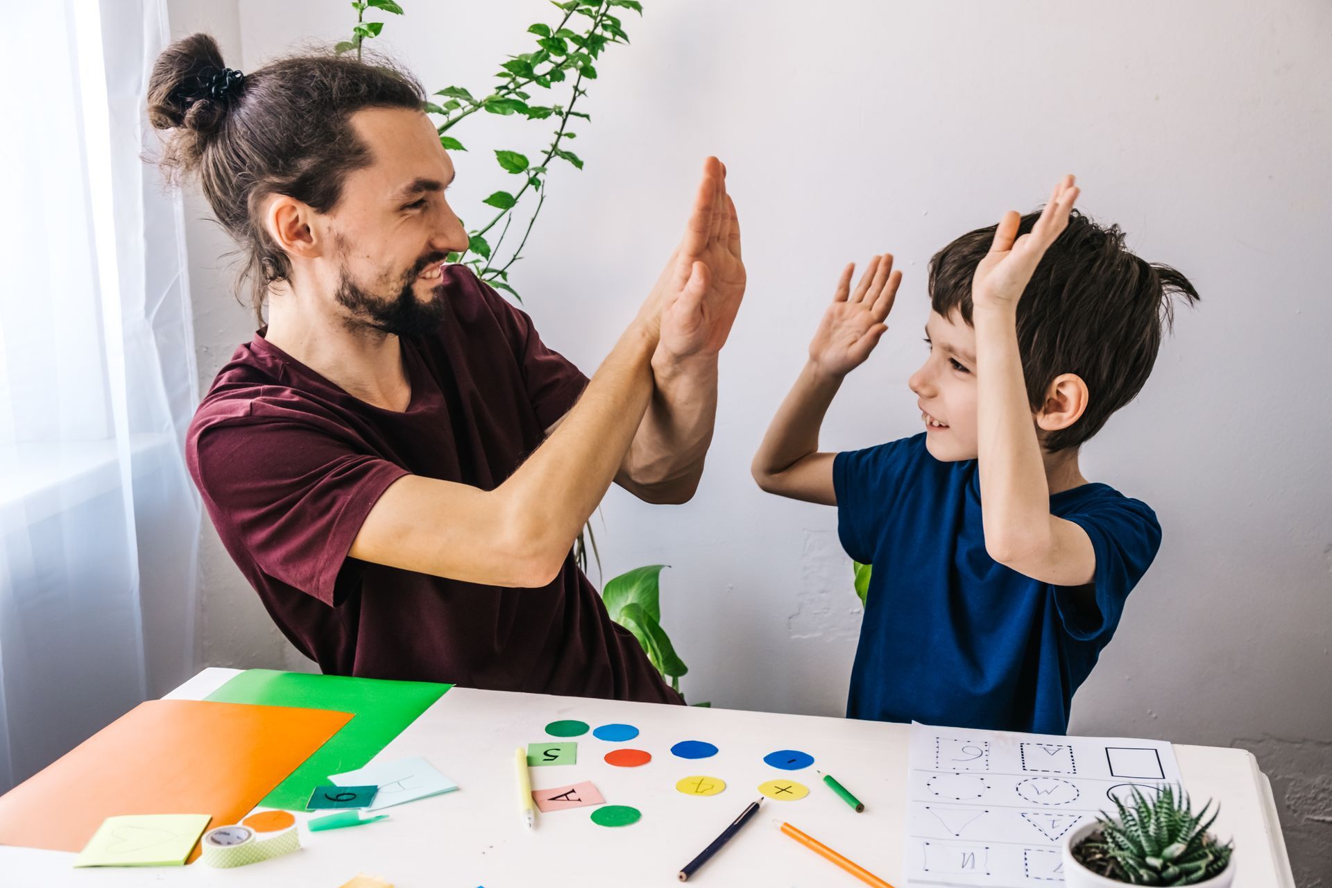 Man and child high-fiving over a table with art supplies. They're smiling in a bright room.