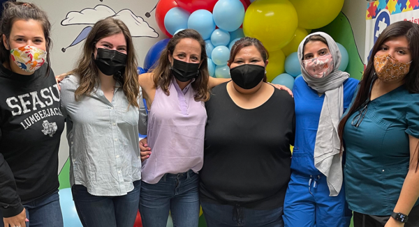 Six people in masks pose together in front of balloons, indoors.